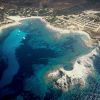 Veduta aerea di una costa con acque blu, barche e terreno arido circostante.