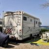 Un camper parcheggiato vicino al mare con sedie a sdraio e tavolino su un tappeto verde.