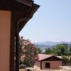 Vista parziale di edifici beige con tetto rosso, alberi e montagne sullo sfondo.